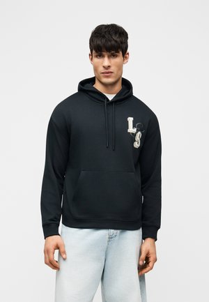 Mann iført svart hettegenser med bokstavene "LS" på brystet og lyseblå jeans, stående mot en ensfarget lys bakgrunn.