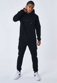 Sudadera negra con bolsillo canguro y cordón, combinada con pantalones cargo que tienen bolsillos laterales. Estilizada con zapatillas Nike blancas.