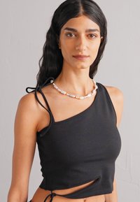 Femme avec de longs cheveux bruns portant un haut noir à une épaule et un collier en perles blanches et roses, se tenant contre un mur clair et uni.