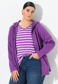 Sudadera con cremallera morada de textura, combinada con una camiseta de cuello en V a rayas morada y blanca. Acompañada de jeans azul oscuro.