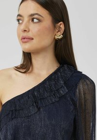 Haut asymétrique bleu marine avec volants, fabriqué en tissu texturé. Présente une manche longue transparente et un éclat métallique contrasté. Boucle d'oreille en forme de cœur doré.