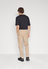 Man in een zwart T-shirt, beige cargobroek en zwarte wandelschoenen, staand tegen een effen lichtgrijze achtergrond, met zijn gezicht naar weg.