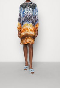Flerfärgad kort klänning med zigzag-mönster, med långa ärmar och hög krage. Bärs med ljusblå sandaler i slid-modell.