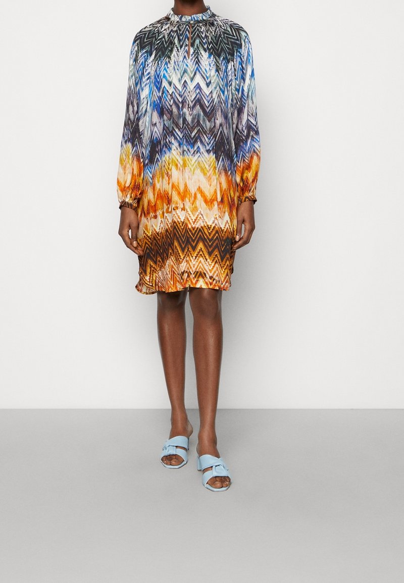 Flerfärgad kort klänning med zigzag-mönster, med långa ärmar och hög krage. Bärs med ljusblå sandaler i slid-modell.