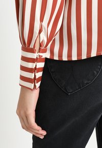 Rød og hvid stribet bluse med en sammensnøret, plisseret ærmedetalje. Stoffet ser glat ud; parret med sorte bukser med en hjerteformet lomme.