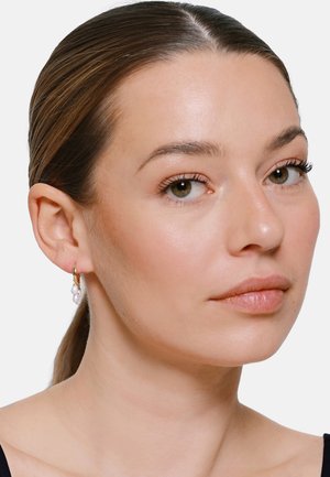 Kobieta o jasnej karnacji i brązowych włosach, nosząca złote kolczyki-koła z wiszącymi białymi perłami, spogląda lekko w prawo, na jednolitym tle.