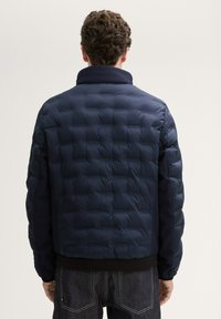 Marineblaue Steppjacke mit einem gesteppten Muster, hohem Kragen, gerippten Bündchen und einem einfarbigen Rückendesign; kombiniert mit dunklen Jeans.