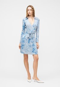 Γυναίκα που φοράει μπλε φόρεμα με λουλούδια, τύπου wrap, με μακριά μανίκια και ζώνη στη μέση, συνδυασμένο με λευκά χαμηλοτάκουνα παπούτσια slingback.