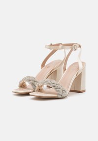 Beige blokk-hælsandaler med en flette, dekorert stropp. Laget av glatt materiale, med ankelremmer og en polstret såle.