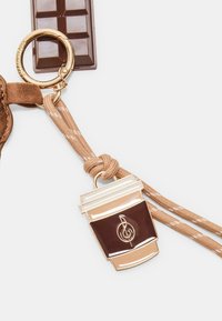 Porte-clés en forme de tasse à café marron avec un motif de note de musique, attaché à une sangle beige et un anneau doré, à côté d'une barre de chocolat.