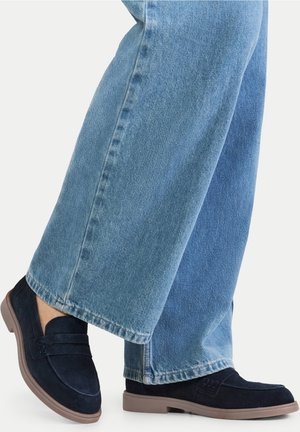 Mocassini in camoscio blu navy con design a profilo basso e tomaia testurizzata. La suola è in gomma marrone, abbinata a jeans a gamba larga di colore azzurro chiaro.