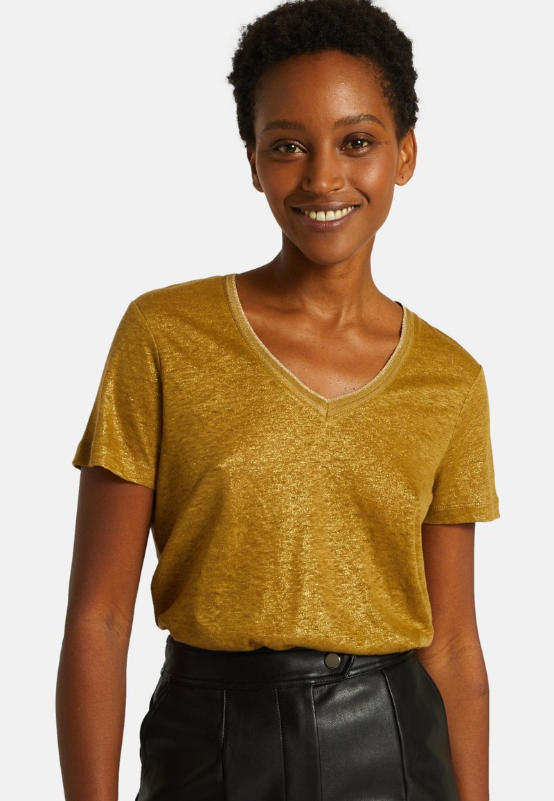 T-shirt doré à manches courtes et col en V avec un léger éclat. Texture douce, coupe décontractée, associé à une jupe en cuir noire taille haute.