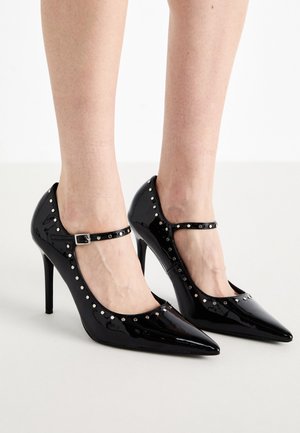 Svarte patentlær høye hæler med spiss tå, ankelstropper og metalliske nagler rundt kantene. Tynne stilettohæler.