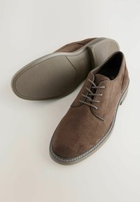Chaussures en daim marron avec un bout rond, un laçage, et une semelle en caoutchouc présentant un motif texturé. Coutures contrastées le long du bord.