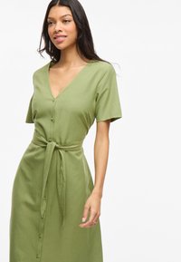 Femme portant une robe boutonnée verte clair avec des manches courtes et une ceinture nouée à la taille, se tenant devant un fond blanc uni.