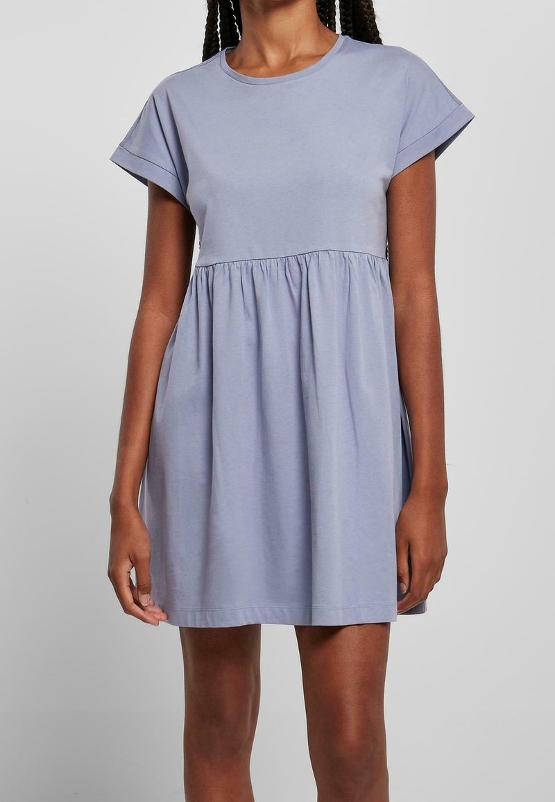 Frau in einem kurzärmligen, hellblauen, knielangen Kleid mit gerafftem Taillendetail vor einem schlichten grauen Hintergrund.