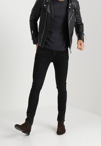 Czarna skórzana kurtka motocyklowa z srebrnymi zamkami, strukturalny ciemny sweter, czarne jeansy slim-fit oraz brązowe zamszowe botki za kostkę.