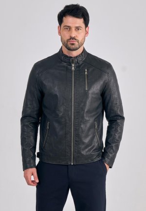 Mężczyzna o ciemnych włosach i brodzie, ubrany w czarną skórzaną kurtkę z zasuwanymi kieszeniami, stojący na tle jednolitego jasnego tła.