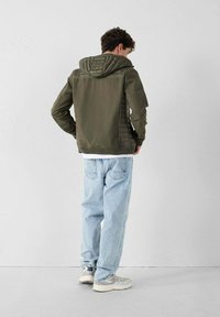 Giacca verde oliva con cappuccio voluminoso e testurizzato, pannelli laterali a contrasto, abbinata a jeans blu chiaro e sneakers grigie.