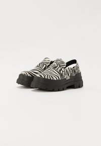 Črno-bela zebra vzorec slip-on čevlji z teksturirano zgornjimi deli iz tkanine, prevelikim črnim gumijastim podplatom in nastavljivim trakom.
