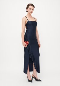Navy satin kjole med tynne stropper, asymmetrisk volangdetalj, sidesplit, kombinert med svarte slingback hæler med gullakcent. Rød hjerteformet clutch.