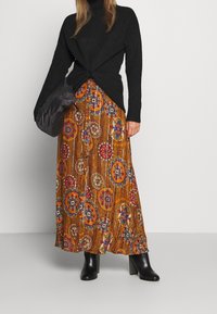 Maglione nero a collo alto in maglia con una parte frontale attorcigliata, abbinato a una lunga gonna a fantasia nei toni dell'arancione, marrone e con motivi floreali multicolori. Stivaletti neri con tacco.