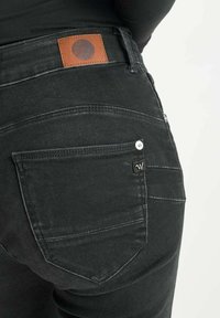 Jean en denim noir avec une pièce en cuir, des coutures visibles et une poche arrière avec un détail de bouton en métal.