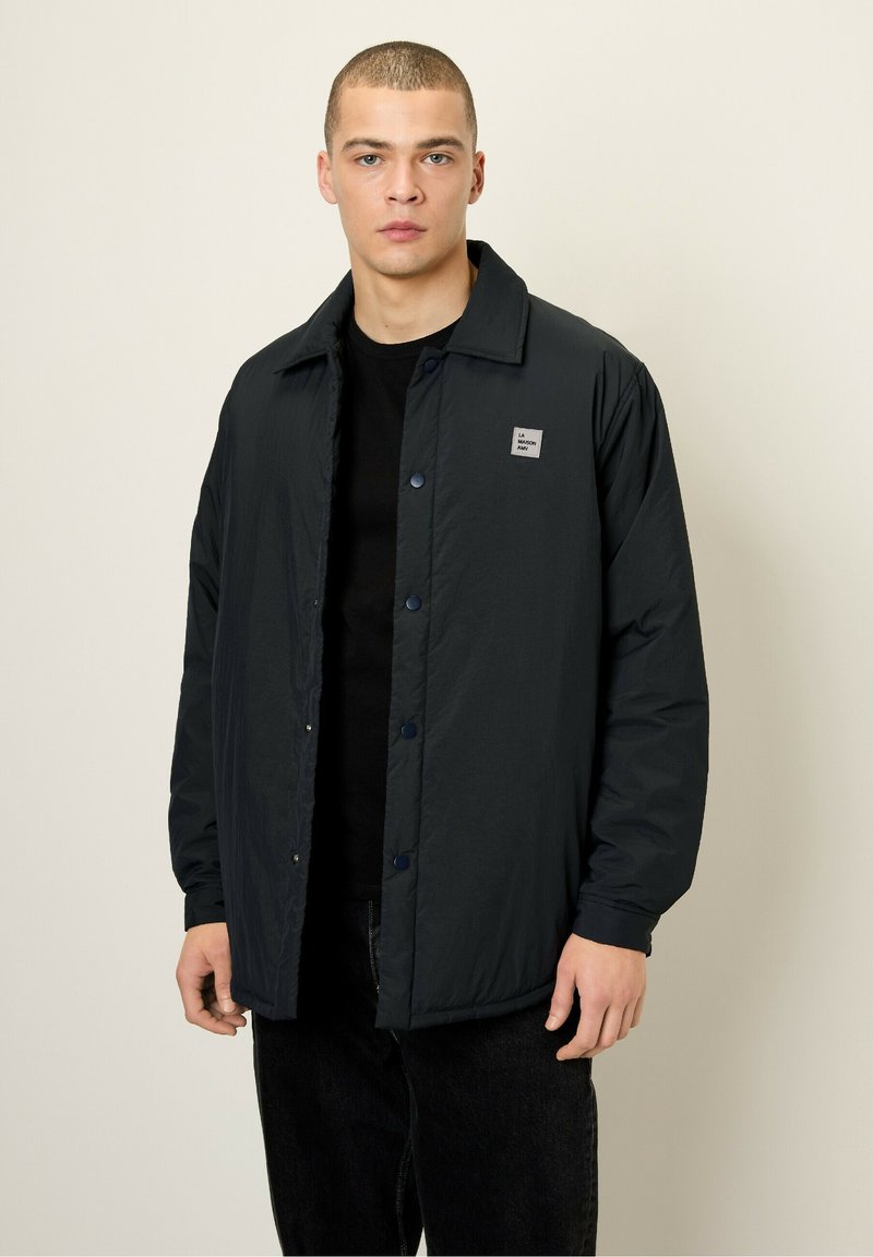 Dark blue nylon jacket with a button front, pointed collar, and a small logo patch on the chest. Features long sleeves and a relaxed fit.