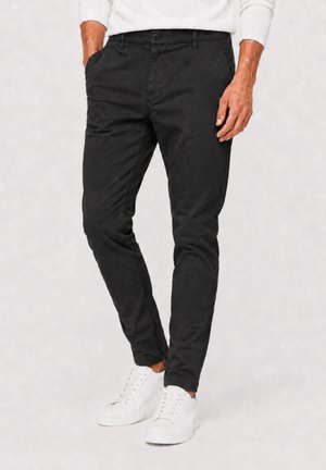Mann trägt schwarze Slim-Fit-Hose mit Händen in den Taschen und weißen Schnürschuhen vor schlichter Hintergrund.