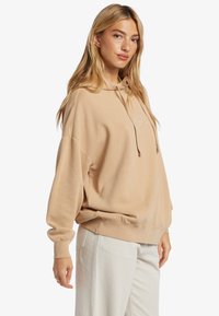 Sudadera con capucha de color beige y corte relajado, con un logo en el frente. Textura suave y puños de canalé. Combinada con pantalones de pana de color claro.