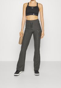 Svart sport-BH med logotyp, grå löst sittande sweatpants med snörning och svarta sneakers. Håller en beige vattenflaska.