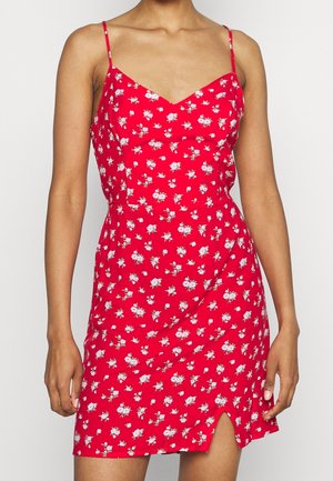 Robe d'été rouge à fleurs avec bretelles fines, décolleté en V et fente sur le côté. Présente de petites fleurs blanches sur un tissu texturé.