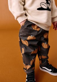 Pantalon gris avec des illustrations de citrouilles orange, assorti à un pull crème et des baskets noires. Taille ajustable et poches latérales visibles.