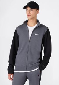 Grau-schwarzer Zip-Hoodie aus Stoff. Verfügt über einen hohen Kragen, elastische Bündchen und Logo-Akzente auf der Brust und dem Ärmel.