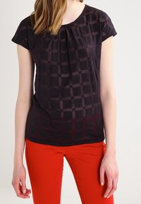 Černý top s texturovaným mřížkovým vzorem, krátkými rukávy a nařaseným výstřihem, spárovaný s úzkými červenými kalhotami.