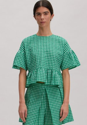 Frau trägt ein grün-weiß kariertes Kurzarm-Top mit Rüschensaum und passenden plissierten Rock, steht vor hellem Hintergrund.