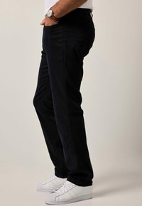 Pantaloni neri slim fit, realizzati in morbido cotone, abbinati a scarpe da ginnastica bianche e un orologio da polso, mostrati di lato.