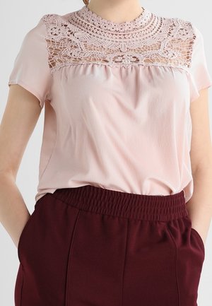 Woman wearing a light pink blouse with intricate lace detail on the upper chest and short sleeves, paired with high-waisted dark red pants.