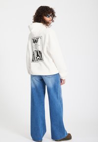 Personne aux cheveux bouclés portant des lunettes de soleil noires, un sweat à capuche blanc avec un motif de soleil noir dans le dos, un jean bleu à jambes larges et des chaussures marron.