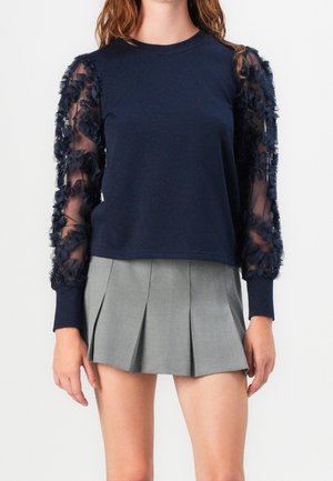 Kobieta w granatowej bluzce z długim rękawem i przezroczystymi rękawami w kwiatowy wzór oraz w szarej plisowanej mini spódnicy.