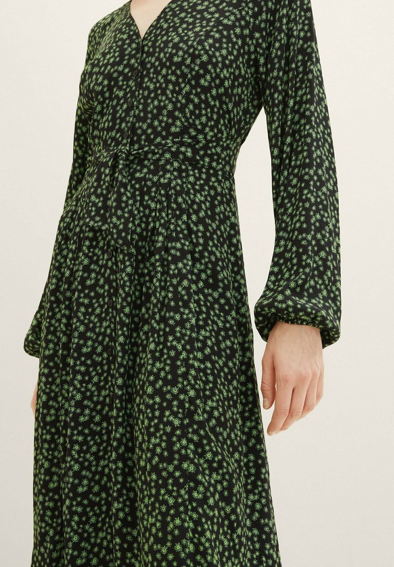 Schwarzes Kleid mit langen Ärmeln, V-Ausschnitt und grünem Blumenmuster. Verfügt über Knöpfe entlang der Vorderseite und ein tailliertes Oberteil mit ausgestellt Rock.