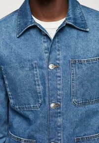 Veste en denim bleu clair avec fermeture à boutons, col évasé et deux poches poitrine ; présente une surface texturée et des coutures visibles.