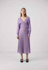 Robe en tricot côtelé lavande avec un col en V, des manches longues et un corsage ajusté qui se transforme en une jupe plissée. Talons aiguilles noirs.