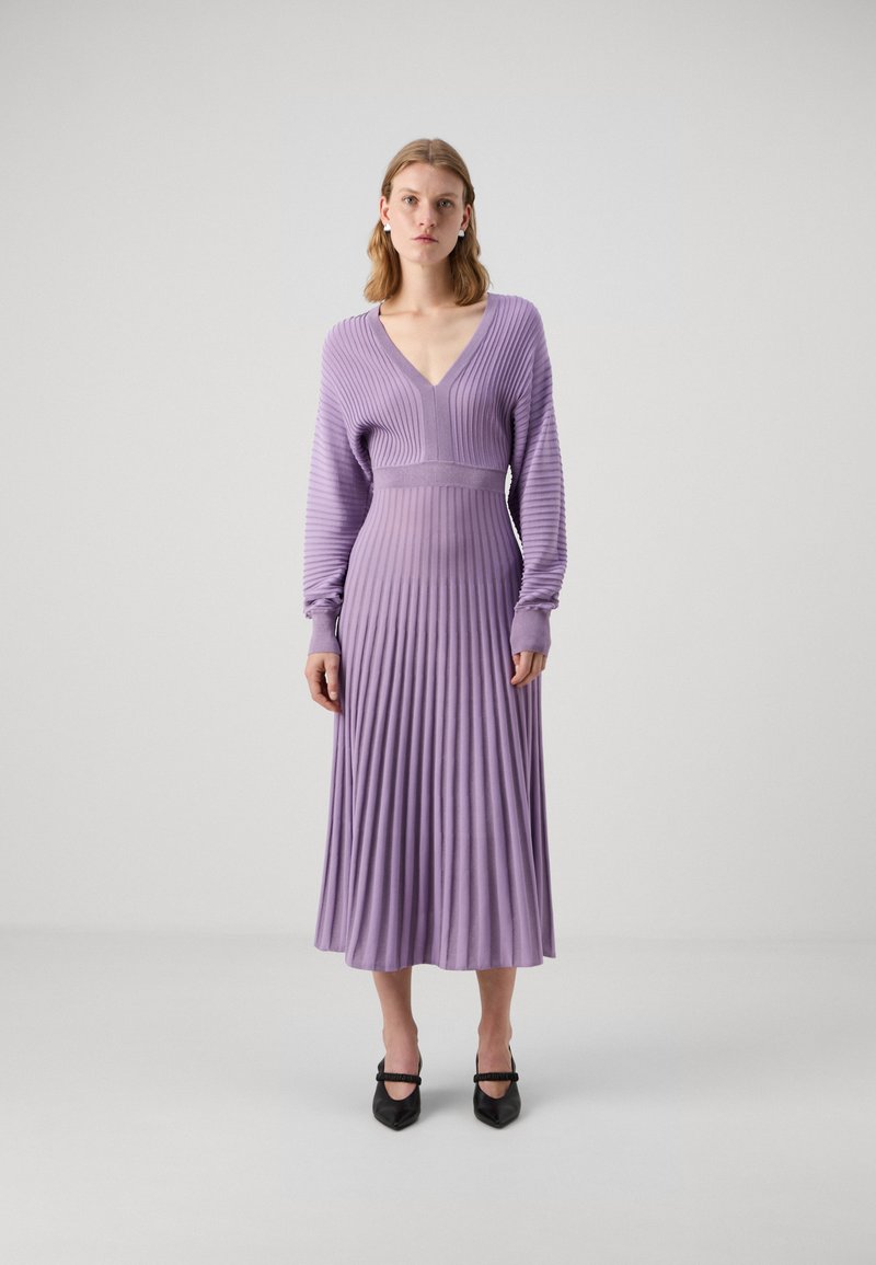 Robe en tricot côtelé lavande avec un col en V, des manches longues et un corsage ajusté qui se transforme en une jupe plissée. Talons aiguilles noirs.