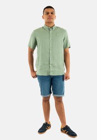 Camicia di lino verde a maniche corte con bottone frontale, abbinata a pantaloni in denim arrotolati e sneaker nere con dettagli arancioni.