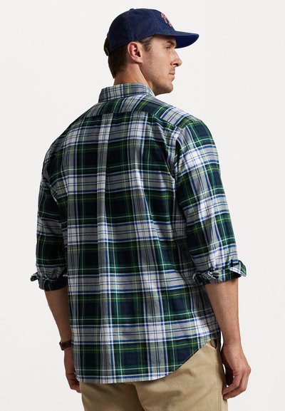 Man met opgerolde mouwen een groen, wit en marineblauwe geruite shirt, beige broek en een marineblauwe baseballcap, van achter en opzij getoond.