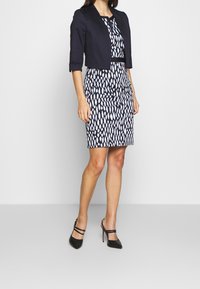 Veste courte navy sur une robe ajustée à motifs noir et blanc. Des escarpins noirs à talons hauts complètent la tenue.