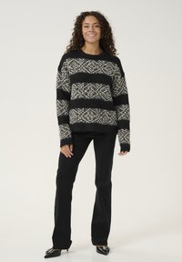 Črno-bež pulover z vzorcem in udobno teksturo, ki ima vodoravne črte. Usklajen z črnimi širokimi hlačami in konicami čevlji.