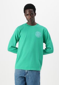 Long-sleeve green cotton t-shirt with a round neckline and a circular logo in white on the left chest. Worn with blue denim jeans.