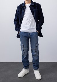 Námořnická manšestrová košile, bílý tričko, modré cargo džíny s kapsami a bílé tenisky. Neformální oblečení s funkčním designem a texturami.